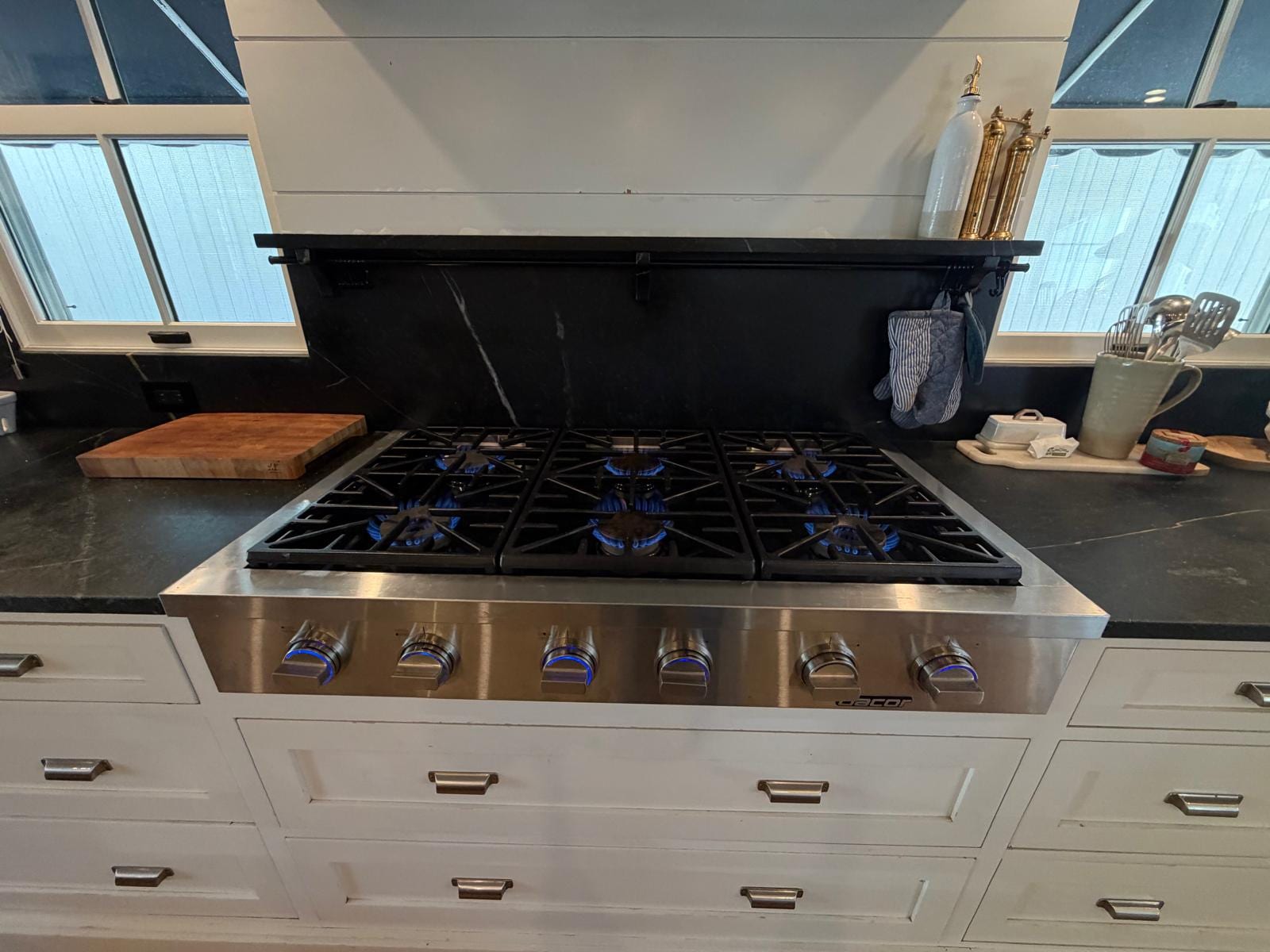 Stainless six-burner Dacor gas rangetop with cast-iron grates, all six burners lit with blue flames and blue-lit control knobs, white cabinets below, a wood cutting board on the left counter and windows on both sides, Newport Beach