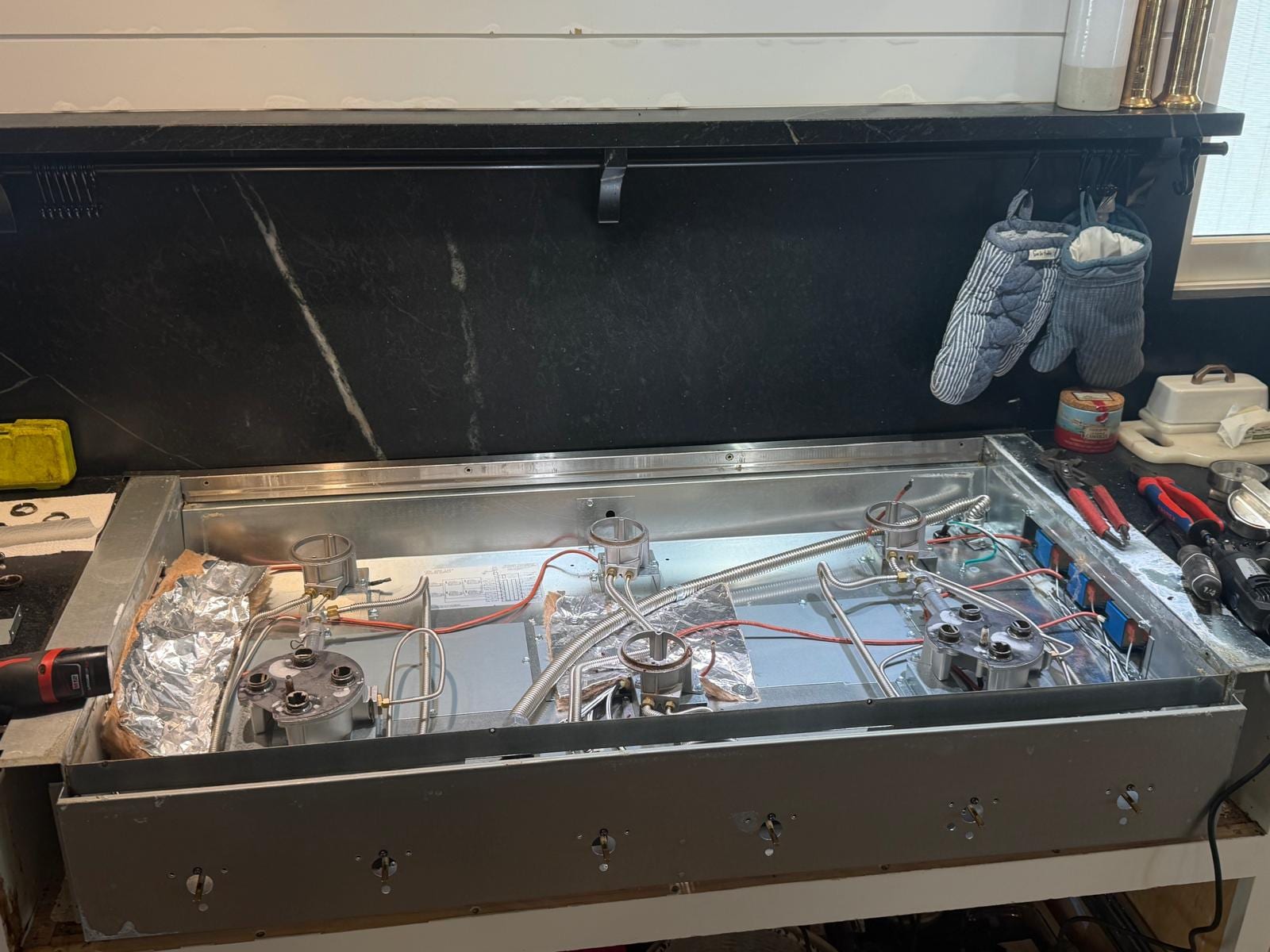 Six-burner gas cooktop built into white cabinetry under a black stone backsplash and shelf in Newport Beach, with the cooktop top removed to show the burner bases, flexible gas lines, red and white wiring and a row of small components along the right side, a cordless drill on the left counter and pliers on the right counter
