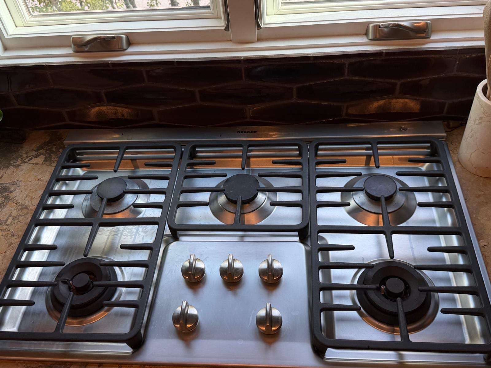 Stainless Miele five-burner gas cooktop with cast-iron grates set into a stone counter below a window in a Corona del Mar kitchen