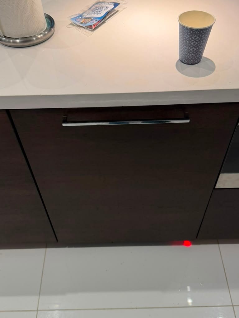 The same Bosch dishwasher closed behind a custom dark wood cabinet panel with a chrome bar handle, red indicator beam still visible on the floor tile