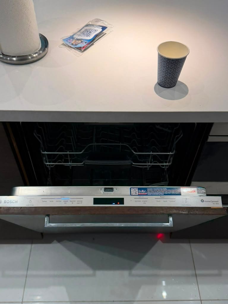Panel-ready Bosch dishwasher with the door open, showing the empty racks and control panel along the top edge of the door, with a red indicator beam on the kitchen floor tile