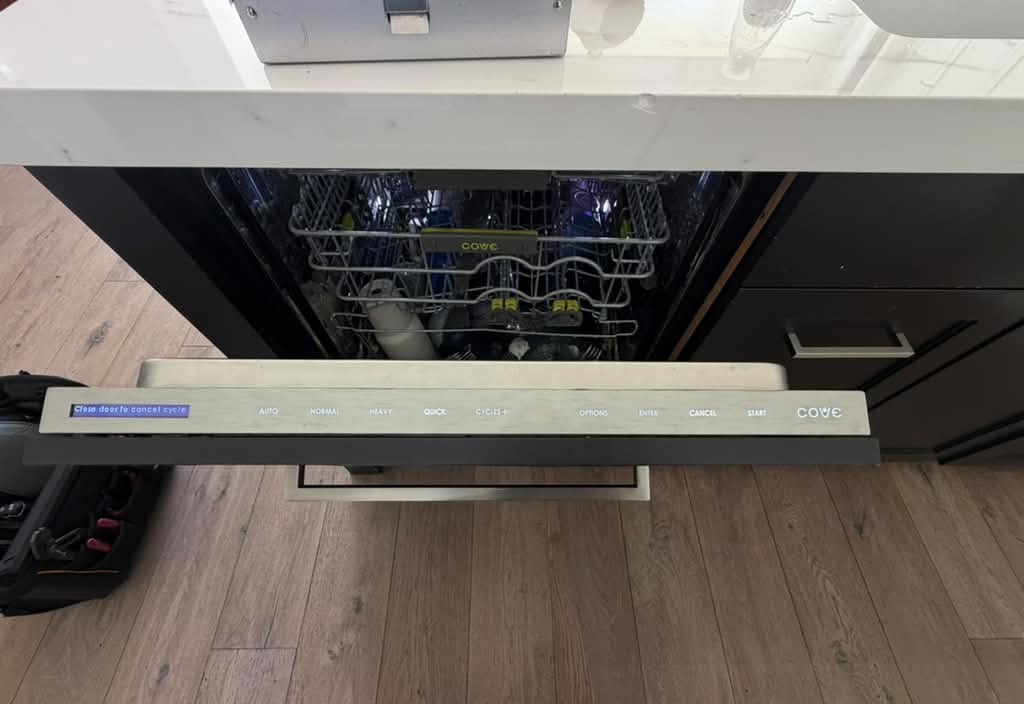 Cove dishwasher with its door partly open, racks loaded and the control strip lit, set in a kitchen island under a white countertop beside dark cabinets in a San Clemente kitchen, with a tool bag on the wood floor