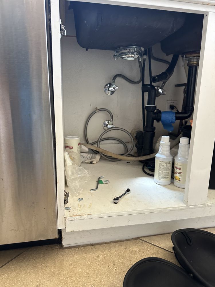 Open kitchen cabinet under a sink showing a garbage disposal mounted below the sink basin, with the drain trap, a coiled dishwasher hose, and a wrench left on the cabinet floor