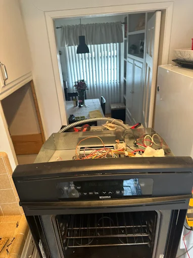 Black built-in wall oven with the top control panel removed, exposing internal wiring, positioned in a kitchen doorway during a repair
