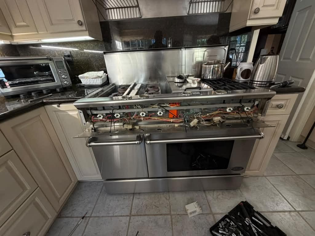 DCS range in an Orange, CA kitchen with cream cabinets and a dark granite backsplash, its front control panel and knobs off with wiring harnesses and valve stems exposed along the front, and an open socket set case on the tile floor