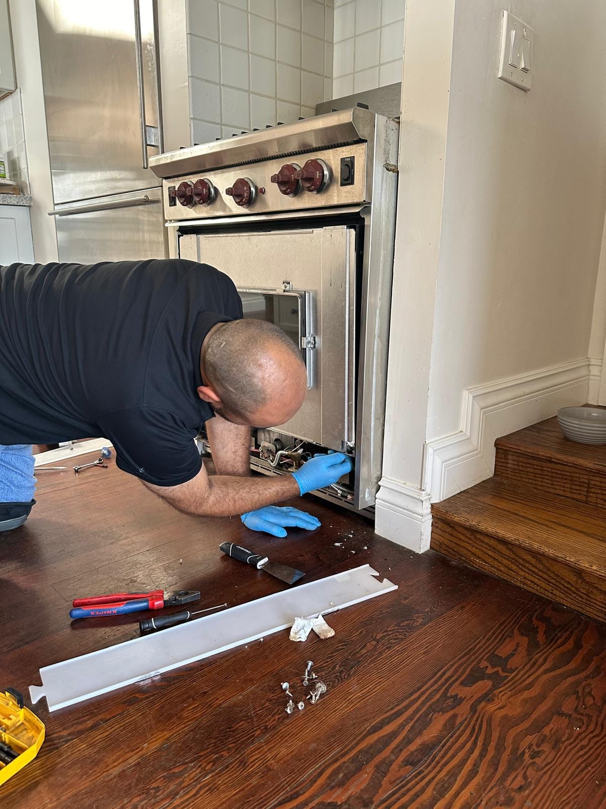 Technician working at the lower front of a stainless Wolf gas range with the trim panel removed and set on the floor, during an oven door hinge replacement in Laguna Beach