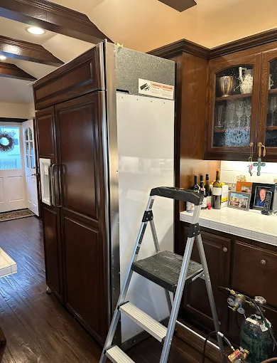 Built-in wood-paneled refrigerator column pulled partway out of its kitchen cabinet enclosure, with a step ladder set up beside it