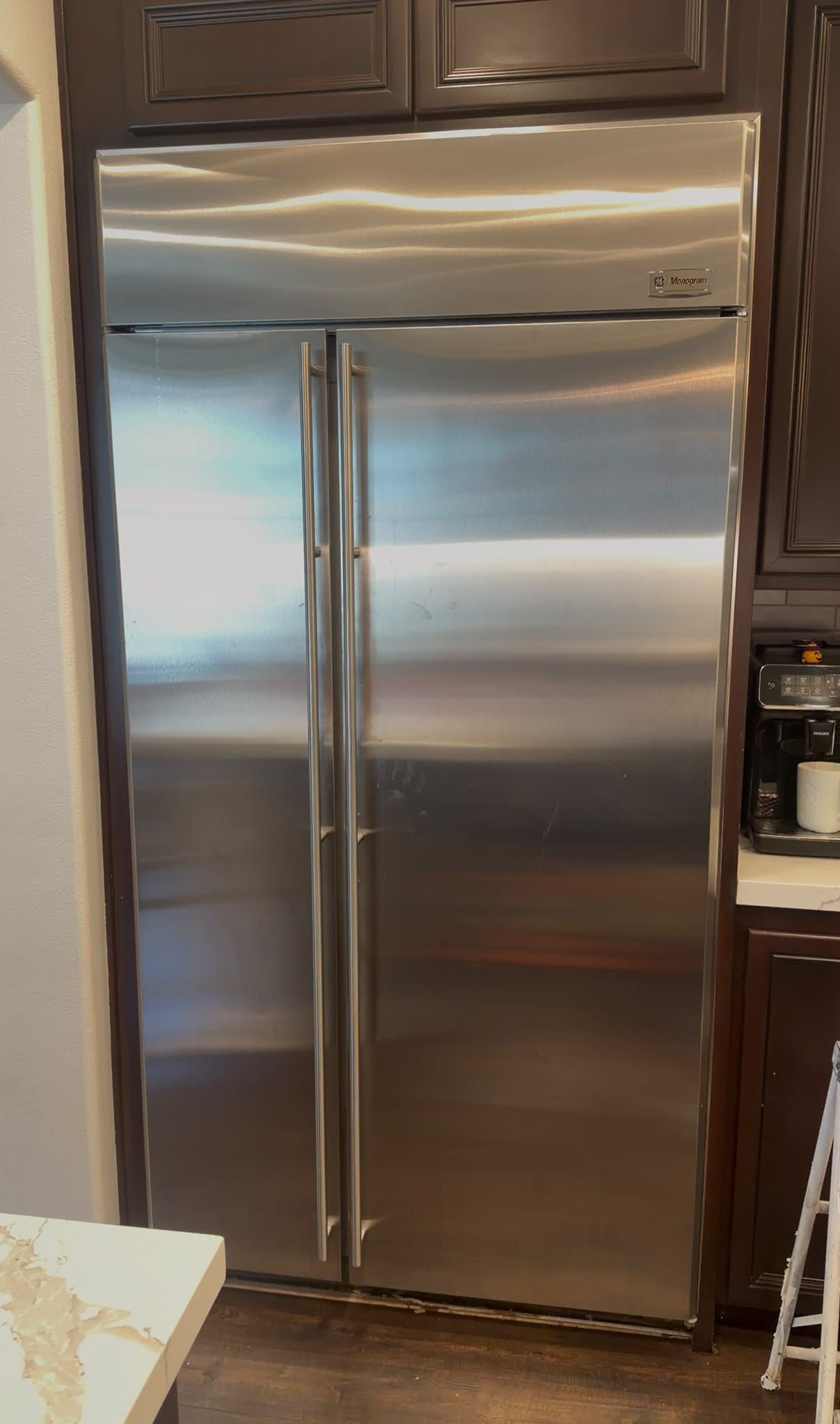 Stainless built-in Monogram refrigerator with its top grille closed beneath brown cabinets
