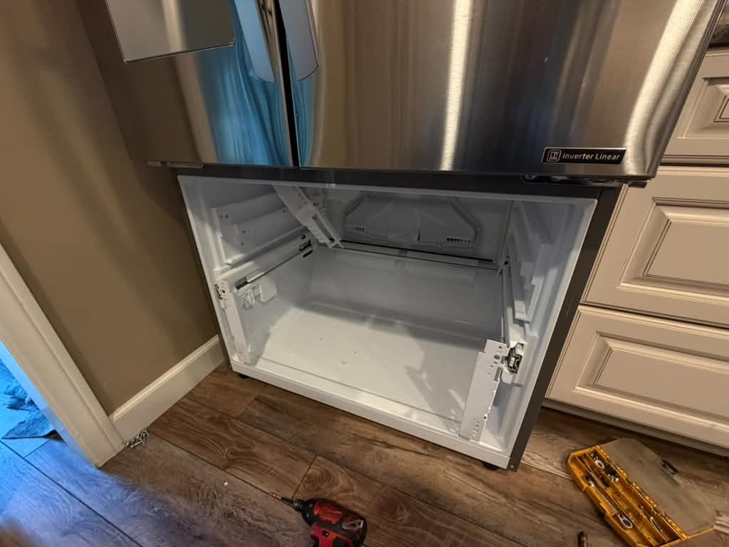 LG refrigerator in Orange, CA with its bottom freezer drawer removed, showing the empty white freezer compartment and drawer rails, with a cordless drill and an open bit case on the floor