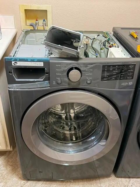 Graphite LG front-load washer in an Orange, CA laundry closet with its top panel removed and the old detergent dispenser housing lifted out, the white replacement assembly resting on top