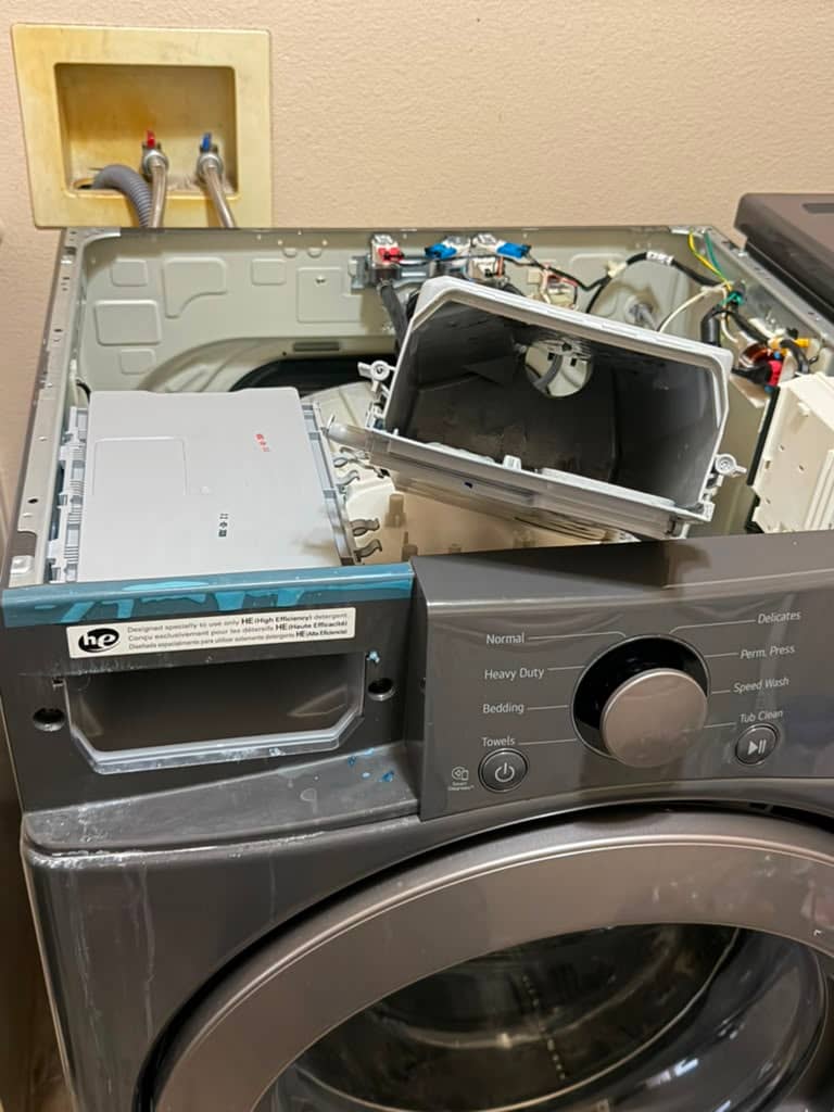 Close view into the open top of an LG front-load washer showing the old detergent dispenser housing tilted out with hardened build-up caked inside, beside the clean white replacement assembly, in Orange, CA