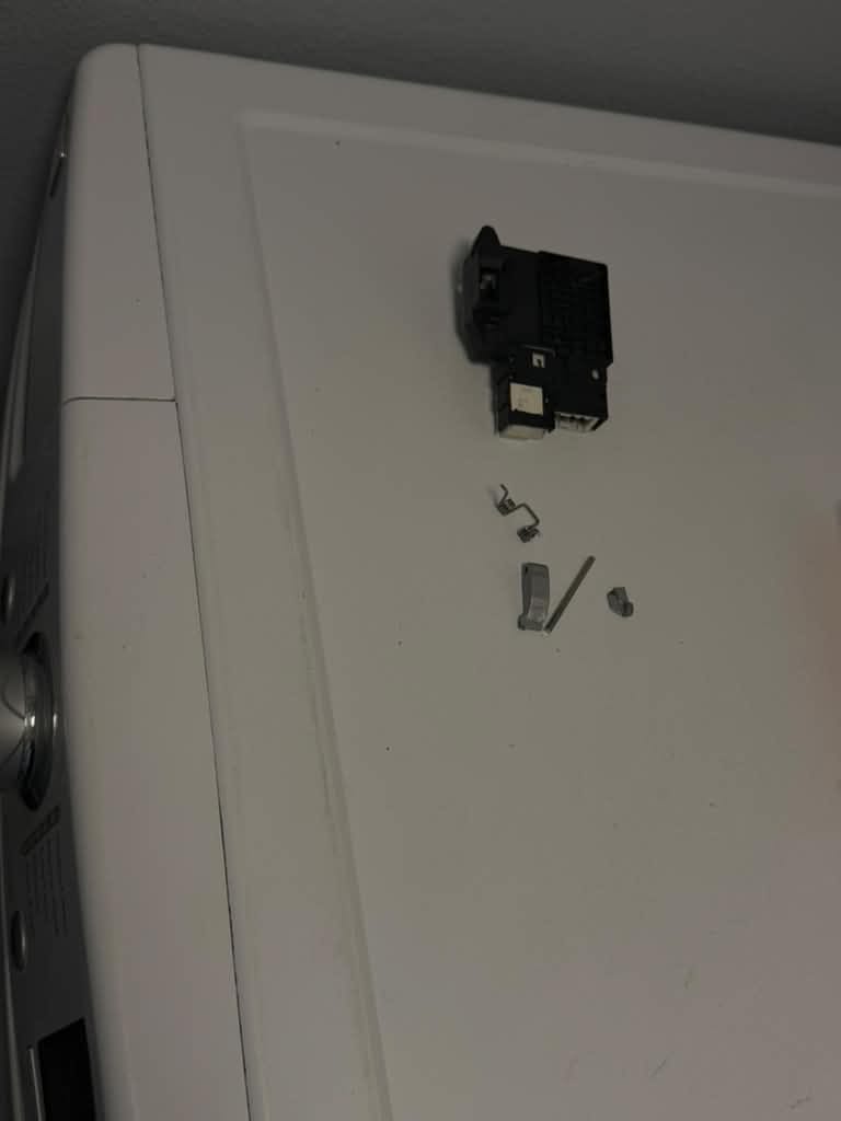 Old parts removed from an LG front-load washer in Cypress, laid out on a white surface: the door lock assembly, a small spring, the door hook and its pin