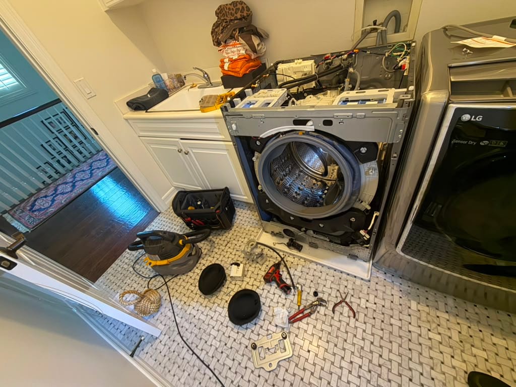 LG front-load washer with the top and control panel removed, exposing the circuit boards, wiring harness, and drum, with tools spread on the tile floor beside a matching LG dryer in a Ladera Ranch laundry room